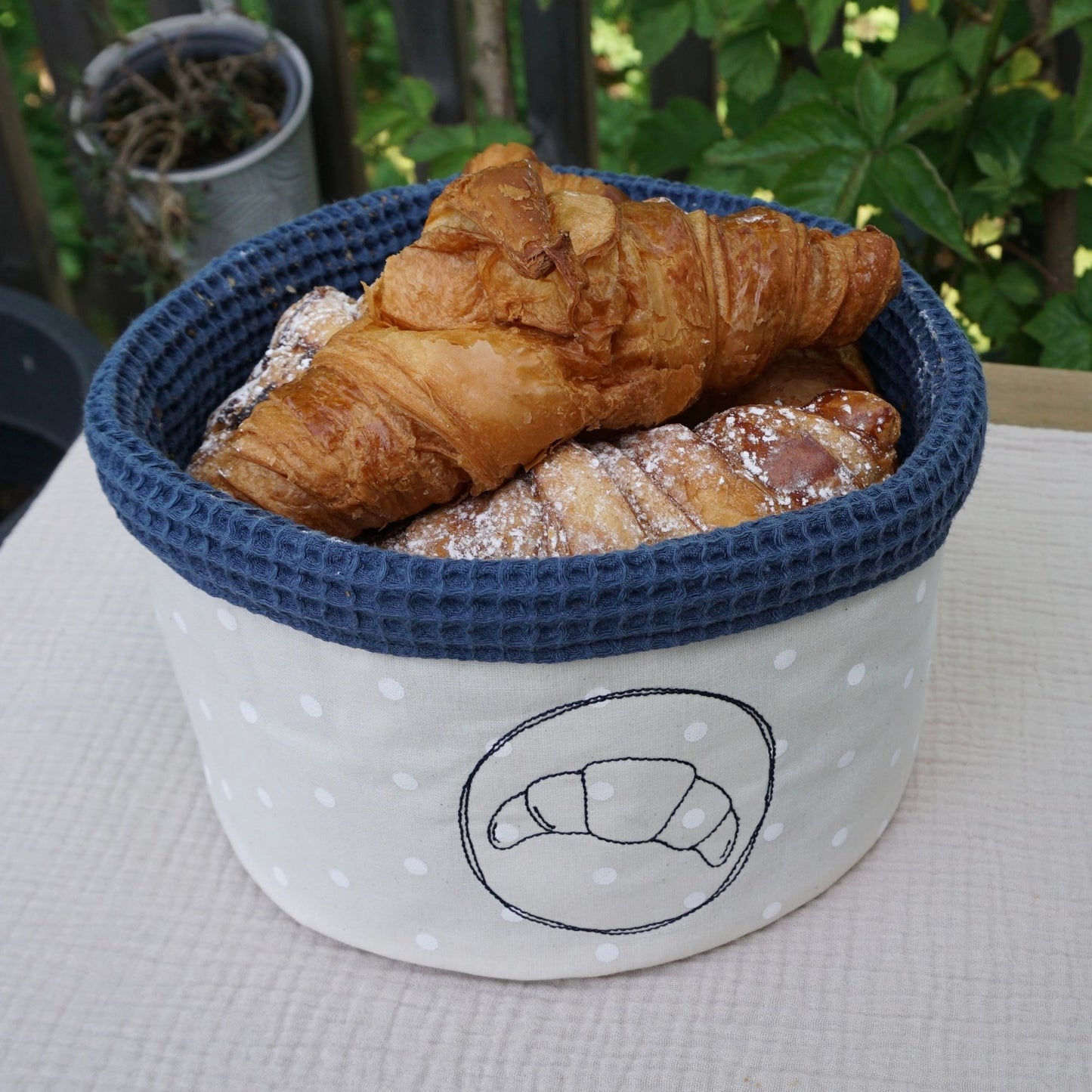 Runder Brotkorb aus Stoff genäht und mit einem Croissant bestickt. Steht gefüllt mit Croissants auf einem Gartentisch.